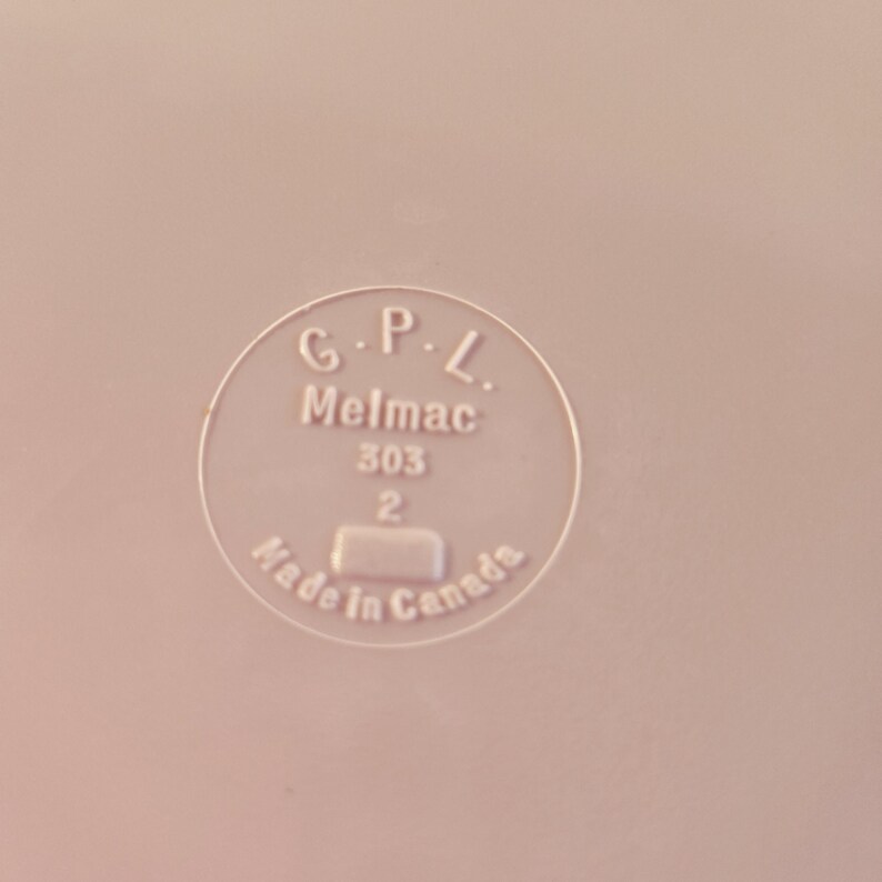 K&ouml;nnte beinhalten: Nahaufnahme einer hellrosa Oberfl&auml;che mit einem kreisf&ouml;rmigen Emblem. Das Emblem lautet "G.P.L. Melmac 303 2 Made in Canada" in einer klaren serifenlosen Schrift. Das Bild ist gut beleuchtet und der Text ist gut lesbar.
