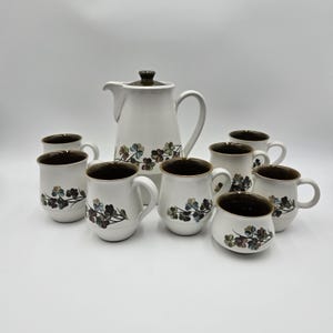 May include: A white ceramic coffee set with a matching coffee pot and mugs. The set features a brown rim and a floral design in shades of green, brown, and blue. The coffee pot has a lid and a handle. The mugs have a handle.