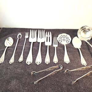 May include: A selection of antique silver serving utensils, including forks, spoons, a ladle, and tongs. The silverware features ornate floral detailing and is arranged on a dark grey surface.