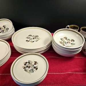 May include: A set of cream-coloured ceramic dinnerware featuring a floral design. The set includes plates, bowls, and serving pieces. The plates and bowls have a light green trim and a floral pattern in brown, blue, and green.