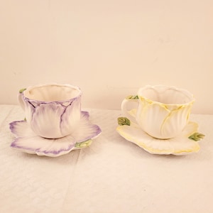 Puede incluir: Dos tazas de té y platillos florales. La taza de té y el platillo de la izquierda son morados y blancos. La taza de té y el platillo de la derecha son amarillos y blancos.