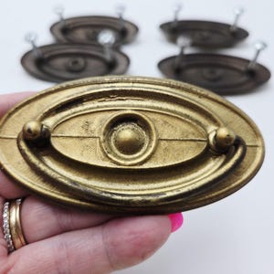 May include: Close-up of a gold-toned oval drawer pull with a central circular design and two small handles. Several similar pulls are visible in the background. The pulls have a vintage, antique appearance.