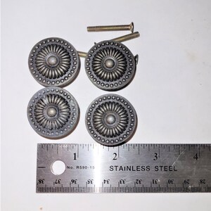 May include: Four round, dark metallic cabinet knobs with a central circular boss surrounded by radiating petals and a beaded outer rim. Two threaded metal screws are included. A stainless steel ruler shows the knobs are approximately 3.8 cm wide.