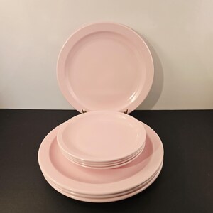 K&ouml;nnte beinhalten: Ein Satz rosa Keramikplatten, darunter gro&szlig;e Essteller und kleinere Teller, gestapelt und auf einer dunklen Oberfl&auml;che angeordnet. Die Teller haben ein schlichtes, klassisches Design mit einem leicht erh&ouml;hten Rand.