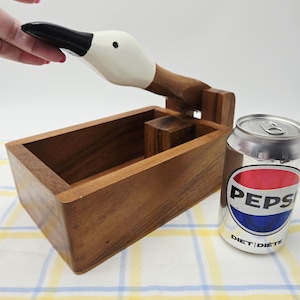 Peut inclure: Un casse-noix en bois en forme d'oie avec une tête blanche et un bec noir. Le mécanisme comporte un cou articulé qui se soulève pour révéler une base en bois rectangulaire. Une canette de soda standard de 330 ml est placée à côté pour l'échelle.