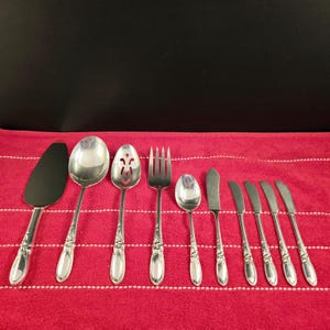 Könnte beinhalten: Ein Set aus zehn versilberten Servierbesteck, bestehend aus einem Kuchenspachtel, zwei Servierlöffeln, einem Schöpflöffel, einer Serviergabel, einem Buttermesser und vier Essmessern. Das Besteck ist auf einer rot-weiß gestreiften Tischdecke angeordnet.