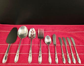 Op de afbeelding: Een set van tien zilverkleurige serveerbestek, waaronder een taartschep, twee serveerscheppen, een geperforeerde lepel, een serveervork, een botermes en vier dinermessen. Het bestek is gerangschikt op een rood-wit gestreept tafelkleed.