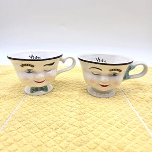 Puede incluir: Dos tazas de cerámica blanca con un rostro esculpido que muestra un ojo guiñando, una boca roja y una pajarita con estampado verde. Cada taza tiene un asa y el texto "Yum." en el borde interior, junto con un pequeño logotipo de Baileys.