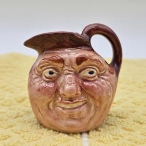 Puede incluir: Una jarra de cerámica con forma de rostro humano y acabado marrón brillante. El recipiente presenta una nariz, ojos y boca sonriente esculpidos, con un asa de color marrón oscuro en el lateral. La superficie tiene una textura lisa y vidriada.