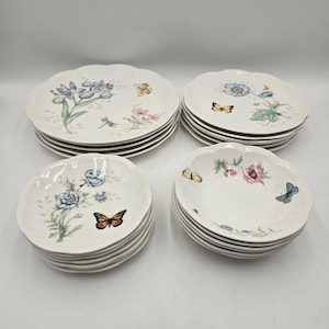 May include: A set of white ceramic plates and bowls with a scalloped edge design. Each piece is decorated with floral and butterfly motifs in blue, pink, and yellow. The plates are stacked, presenting a delicate, nature-inspired aesthetic.