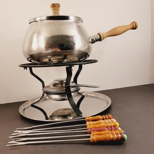 May include: A stainless steel fondue pot with a wooden handle and lid, on a black metal stand with a burner. Includes a silver serving tray and six fondue forks with wooden handles and coloured tips.
