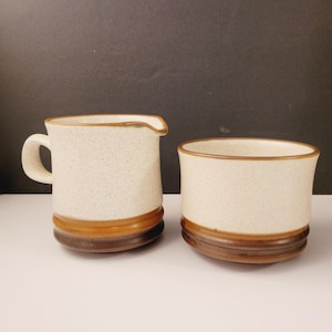 Może przedstawiać: Zestaw dzbanka do śmietanki i cukiernicy z białej ceramiki z brązowymi paskami. Dzbanek ma uchwyt i dzióbek. Cukiernica jest okrągła.