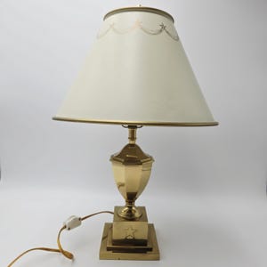 May include: A brass table lamp with a cream lampshade. The lampshade has a gold trim and a star pattern. The base of the lamp is square with a star design. The lamp is plugged into an electrical outlet.