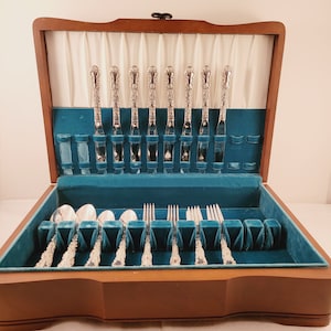 May include: A wooden box with a teal velvet interior, containing a set of silver cutlery. The cutlery is arranged in rows, with the handles facing up. The box is open and the cutlery is visible.