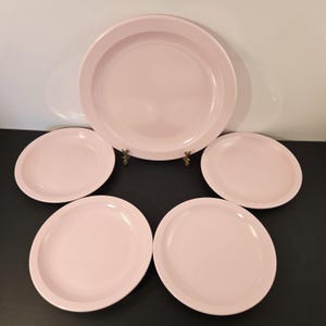 K&ouml;nnte beinhalten: Ein Set aus f&uuml;nf hellrosa Tellern. Das Set besteht aus einem gro&szlig;en Teller und vier kleineren Tellern. Die Teller haben ein schlichtes, klassisches Design mit einem leicht erh&ouml;hten Rand. Die Teller sind auf einer dunklen Oberfl&auml;che angeordnet.