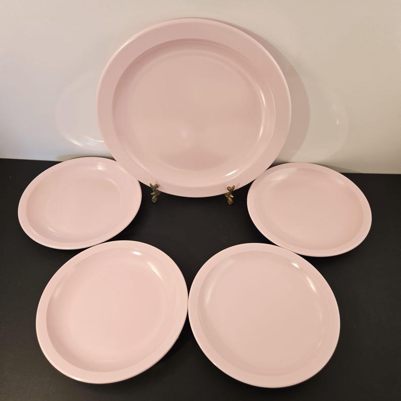 K&ouml;nnte beinhalten: Ein Set aus f&uuml;nf hellrosa Tellern. Das Set besteht aus einem gro&szlig;en Teller und vier kleineren Tellern. Die Teller haben ein schlichtes, klassisches Design mit einem leicht erh&ouml;hten Rand. Die Teller sind auf einer dunklen Oberfl&auml;che angeordnet.