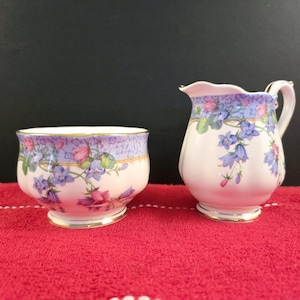 May include: A white sugar bowl and creamer set with a floral pattern featuring pink and blue flowers. The set has a gold trim around the edges.