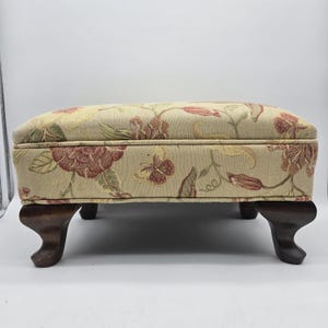 May include: A rectangular footstool featuring a beige fabric upholstery with a woven floral and butterfly pattern in shades of red, green, and yellow. The piece is supported by four dark brown wooden legs with a curved, cabriole-style design.
