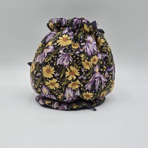 May include: A round, drawstring bag with a floral pattern. The fabric features yellow daisies and purple irises on a dark background. The bag is gathered at the top with a dark blue drawstring.