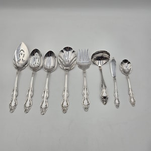 May include: A set of silver-coloured serving utensils, including spoons, forks, and a butter knife. The set features detailed floral patterns on the handles and various serving heads. The utensils are arranged on a light grey surface.