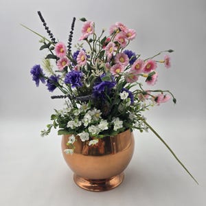 May include: A copper-coloured, round vase filled with an arrangement of artificial flowers. The flowers include pink, purple, and white blooms, with green foliage. The vase has a polished finish and a pedestal base, creating a decorative centerpiece.
