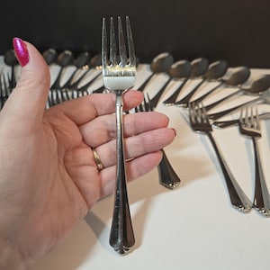 May include: A silver fork with four prongs, held in a hand. The fork has a long, tapered handle with a decorative design at the base. Other silverware, including spoons and forks, are visible in the background, suggesting a set.