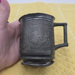 Może przedstawiać: Ciemnoszary metalowy kubek z misternym, tłoczonym wzorem kwiatowym i ornamentami roślinnymi na zewnętrznej powierzchni. Kubek ma cylindryczny korpus z rozszerzanym brzegiem oraz prostokątne ucho o segmentowej konstrukcji przypominającej bambus.