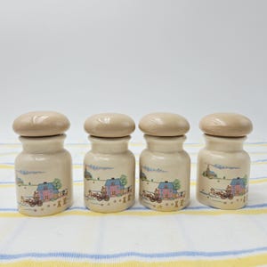 Pode incluir: Um conjunto de quatro potes de cerâmica na cor creme com tampas arredondadas correspondentes. Cada pote apresenta uma cena impressa de uma casa rosa, uma carruagem puxada por cavalos e uma torre de igreja em uma paisagem com árvores.