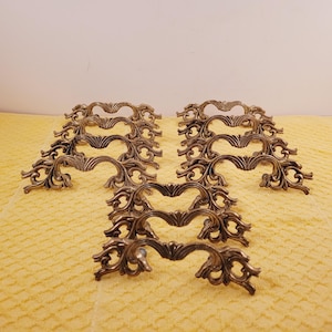 May include: Set of ten ornate brass drawer pulls with a decorative scroll design. The pulls are arranged in rows on a yellow textured surface.