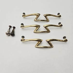 Könnte beinhalten: Drei goldfarbene Metall-Schubladengriffe mit einem markanten Zickzack-Design in der Mitte und zwei separate kleine Metallknöpfe mit Gewindeschrauben. Jeder Griff hat eine geschwungene, längliche Form, die zu den Befestigungspunkten hin schmaler wird.