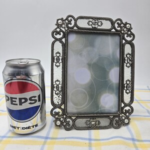 May include: A rectangular metal picture frame with an ornate, dark grey filigree border featuring small white bead accents. The frame surrounds a glass pane with a soft-focus circular pattern. A silver Diet Pepsi soda can is placed beside it.