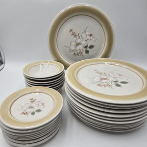 May include: A collection of vintage dinnerware featuring a floral design. The set includes dinner plates, salad plates, and bowls. Each piece has a cream-colored center with a floral pattern and a tan border. The plates and bowls are stacked.