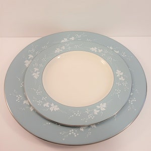 Puede incluir: Un juego de dos platos de cerámica. El plato más grande es azul claro con un estampado floral blanco y un borde plateado. El plato más pequeño es blanco en el centro con un borde azul claro, estampado floral blanco y borde plateado. Los platos están apilados.