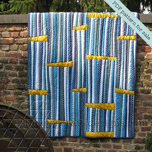 Op de afbeelding: Een quilt met verticale strepen in tinten blauw, grijs en wit, met gele horizontale strepen over de quilt. De quilt hangt aan een bakstenen muur. De tekst "PDF pattern for sale" staat in de rechterbovenhoek van de afbeelding.