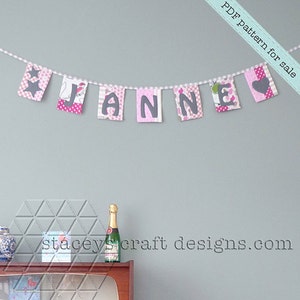 Op de afbeelding: Een roze en grijze stoffen banner met de naam "JANNE" gespeld in grijze letters. De banner hangt aan een lichtblauwe muur. De tekst "PDF pattern for sale" staat in wit op een witte achtergrond in de rechterbovenhoek van de afbeelding. De tekst "staceyscraftdesigns.com" staat in zwart op een witte achtergrond in het midden onderaan de afbeelding.