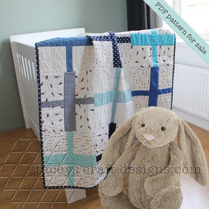 Op de afbeelding: Een blauwe en witte patchwork babydeken met een witte stippenrand. De deken is over een wit wiegje gelegd en een knuffelkonijn zit er voor. De tekst "PDF pattern for sale" staat in de rechterbovenhoek van de afbeelding.