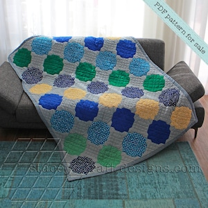 Op de afbeelding: Een grijs, blauw, groen en geel quilt met een geometrisch patroon. De quilt is gevouwen en gedrapeerd over een grijze bank. De tekst "PDF pattern for sale" staat in de rechterbovenhoek van de afbeelding. De tekst "staceycraftdesigns.com" staat in de rechterbenedenhoek van de afbeelding.