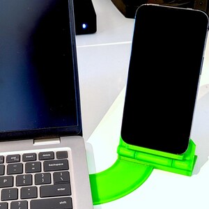 Könnte beinhalten: Ein Laptop und ein Smartphone liegen auf einer weißen Oberfläche. Das Smartphone steht in einem hellgrünen Ständer. Der Laptop ist geöffnet, die Tastatur ist sichtbar. Der Bildschirm des Smartphones ist schwarz.