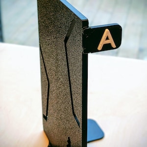 Può includere: Un espositore nero e strutturato con la lettera "A" color oro attaccata. Il supporto ha una forma rettangolare con una base per la stabilità. La superficie ha un aspetto maculato.