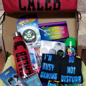 May include: A red gift box with the name "Caleb" printed on the lid. The box is filled with various items, including a water bottle, socks, a yo-yo, a notebook, and candy. The socks have the phrases "I'm Busy Gaming" and "Do Not Disturb" printed on them.