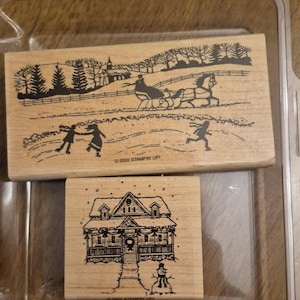 May include: Two rectangular wooden rubber stamps with black ink designs. The larger stamp depicts a winter landscape with a horse-drawn sleigh, trees, and figures skating. The smaller stamp shows a house with a snowman. Both feature 2002 STAMPIN UP text.