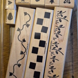 May include: A collection of wooden stamps with various designs, including a heart, flower, swirl, star, and butterfly. The set also includes stamps with vine, square, and leaf patterns. The stamps are stored in a clear plastic container. The text "©2002 STAMPIN' UP!" is visible.