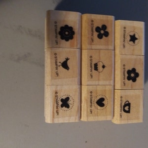May include: Nine wooden stamps with various black designs, including flowers, a bird, a star, a cupcake, a butterfly, a heart, and a cup. Each stamp has the text "STAMPIN' UP!" printed on it.