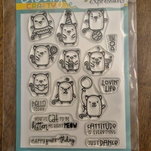 May include: Clear stamp set featuring cartoon cat illustrations with various expressions and accessories. Includes phrases like "Hello Today," "Cattitude is Everything," and "Happy Purr-thday." The set is packaged in a clear plastic sleeve.