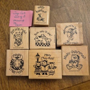 May include: A collection of seven wooden rubber stamps featuring holiday illustrations and text including Merry Kissmas, Just for Mew, From Moo to Ewe, Mistle-Toad, Merry Chris-Moose, Merry Chris-mice, and Beary Christmas, with a pink note.