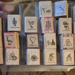 May include: A collection of twenty square wooden rubber stamps arranged in a clear plastic tray. Each stamp features a unique black ink illustration, including a heart, a bee, a king, a clock, a tank, a saw, a weight, and various other symbols.