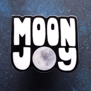 Pode incluir: Um autocolante a preto e branco com as palavras "MOON JOY" em letras arrojadas e arredondadas. Uma imagem detalhada da lua está no centro. O autocolante está sobre um fundo azul escuro com manchas brancas, assemelhando-se a um céu estrelado.