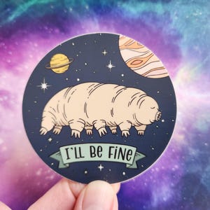 May include: A round sticker with a dark blue background and a cartoon illustration of a tardigrade in space. The tardigrade is pink and has eight legs. The sticker has the text "I'll be fine" in a green banner.