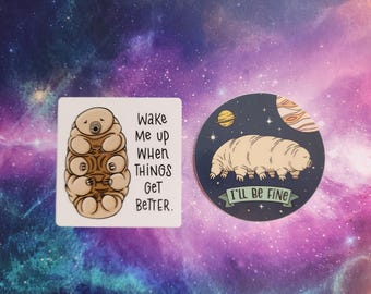 May include: Two stickers featuring a cartoon tardigrade. The first sticker is white with a brown tardigrade curled up and the text "Wake me up when things get better." The second sticker is round with a blue background and a white tardigrade with the text "I'll be fine."