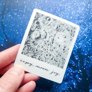 Peut inclure: Un autocollant rectangulaire en forme de photo instantanée vintage présentant une surface lunaire détaillée en noir et blanc avec des cratères. La bordure blanche en bas contient le texte cursif noir qui indique, copy, moon joy.